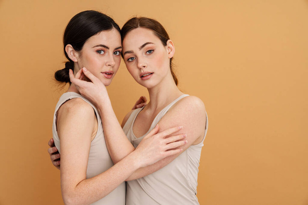 Young two women in undershirt hugging and looking at camera isolated over yellow background - Fotoğraf, Görsel