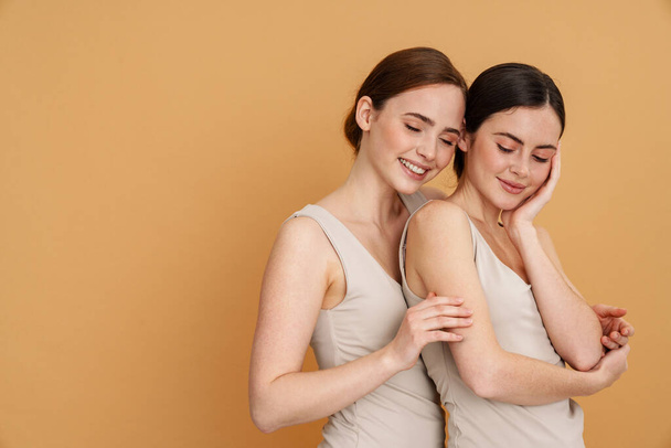 Young two women in undershirt hugging and smiling at camera isolated over yellow background - Foto, imagen