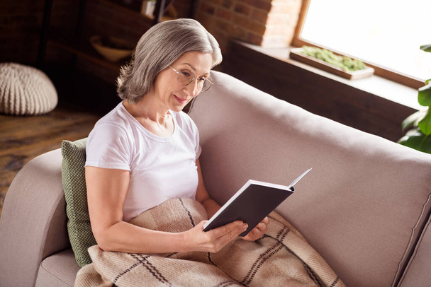 Portrait of attractive focused grey-haired woman sitting on sofa reading interesting book at home house indoors modern brick loft interior - Photo, Image