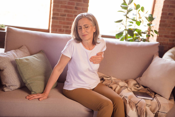 Photo of sad depressed retired woman dressed white t-shirt sitting couch having heart pain arm hand chest indoors flat home house - Foto, Imagem