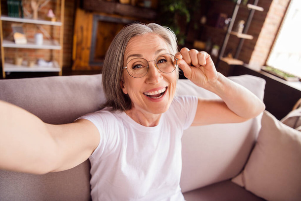 Fotografie hezká roztomilá dáma důchodce nosit bílé tričko sedí pohovka s úsměvem tacking selfie vnitřní dům byt - Fotografie, Obrázek
