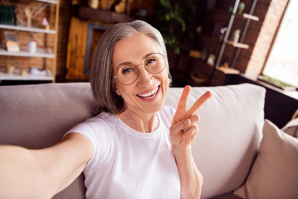 Self-portrait of attractive cheerful grey-haired woman sitting on sofa showing v-sign having fun at home house indoors modern brick interior - Foto, Imagem