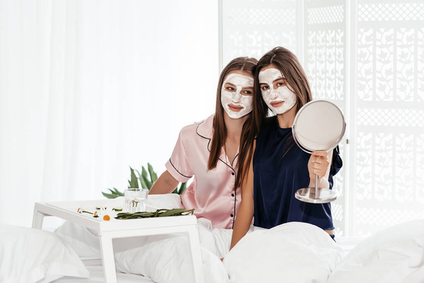 Νεαρές γυναίκες φτιάχνουν πήλινη μάσκα προσώπου κοντά στον καθρέφτη. Κορίτσι που κάνει θεραπεία ομορφιάς και χαλάρωση στο σπίτι. Γυναίκα τεμπέλης κορίτσι χαλάρωση lazing στην κρεβατοκάμαρα. - Φωτογραφία, εικόνα