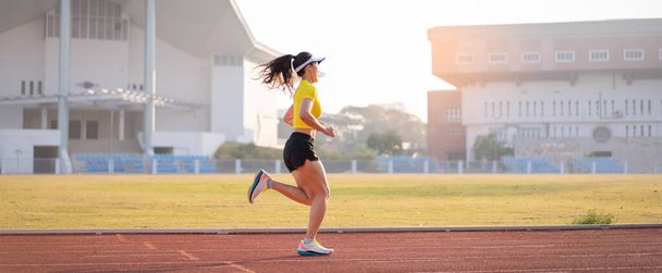 Una joven atleta asiática corriendo en pista de atletismo en el estadio de la ciudad en la mañana soleada para mantener la aptitud y un estilo de vida saludable. Joven mujer de fitness corre en pista de estadio. Deporte y recreación - Foto, Imagen