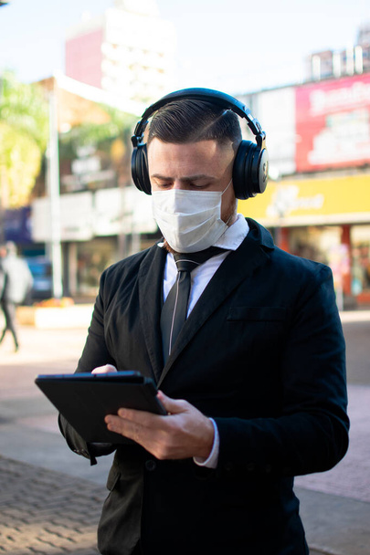 A young male in an elegant suit, medical face mask, and big headphones using a tablet - Фото, зображення