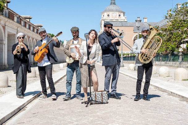 Jazzová kapela hrající na ulici za denního světla a oblečená v bundách, čepicích a šatech, složená ze 6 komponentů, zpěvačky, klarinetu, tuby, trombónu, kytary a podložky. S pozadím zahrad Aranjuez, Madrid, Španělsko. - Fotografie, Obrázek
