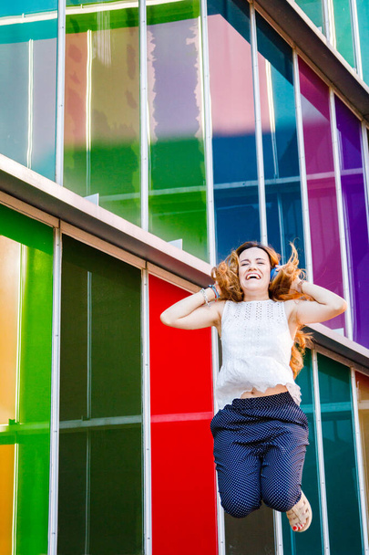 Red head young woman jumping and listening to music over multicolor background - Foto, Imagem
