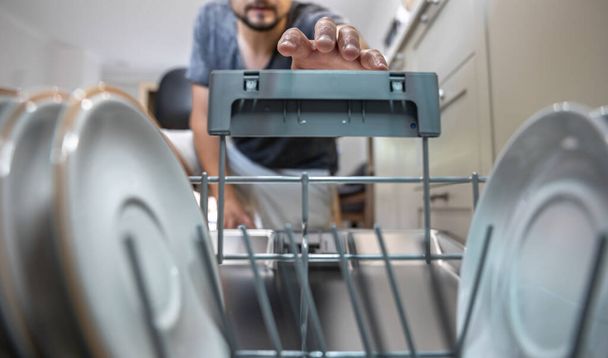 A man in front of an open dishwasher takes out clean dishes after washing. - Foto, Bild
