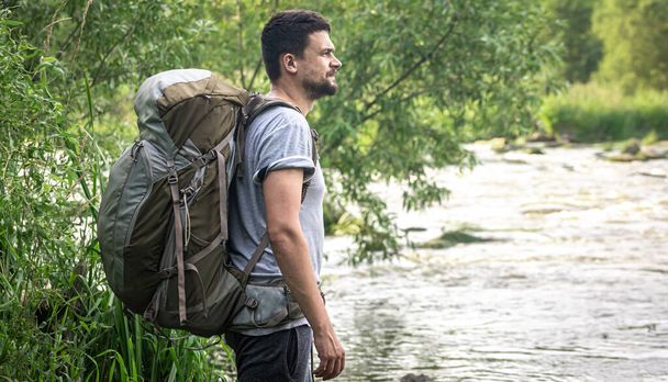 Um viajante do sexo masculino com uma grande mochila de caminhada fica perto do rio. - Foto, Imagem