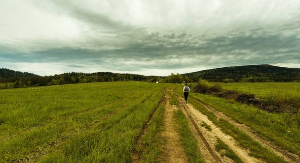 ポーランド・ラッカ・ズドロジ2021年4月10日:ラッカ近郊の丘陵地帯を背景に、バックパックハイキングや牧草地の真ん中を歩く人のパノラマビュー - 写真・画像