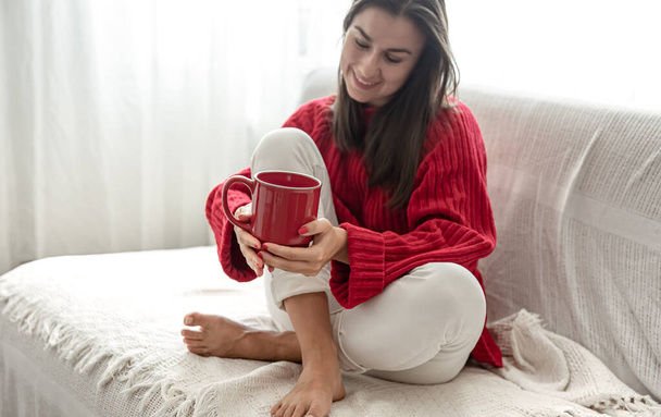 Egy fiatal nő egy piros csésze forró itallal egy kényelmes piros pulóverben otthon pihen a kanapén másolja a helyet.. - Fotó, kép