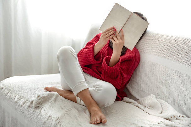 Een vrouw in een rode trui leest een boek terwijl ze op de bank zit. - Foto, afbeelding