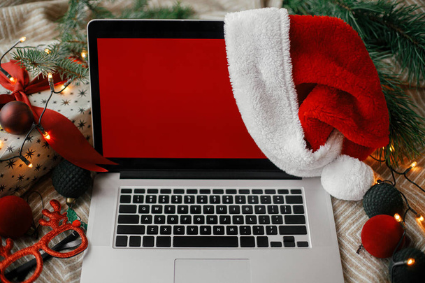 Noel satışları ve online alışveriş. Noel Baba şapkalı, kırmızı boş ekranlı dizüstü bilgisayar şık Noel süsleri, hediyeler ve festival ışıkları olan rahat bir yatakta. Mesaj için yer var. Yukarıya bak. Bağımsız - Fotoğraf, Görsel