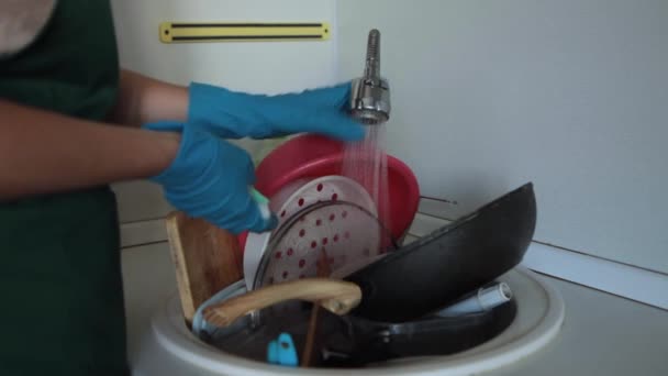 Μια γυναίκα με ποδιά και λαστιχένια γάντια πλένει βρώμικα πιάτα στο νεροχύτη της κουζίνας της. Βρώμικα πιάτα στο νεροχύτη για πλύσιμο. - Πλάνα, βίντεο