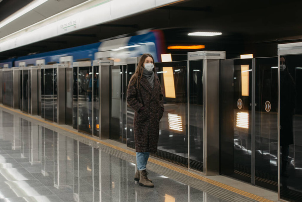 A full-length photo of a woman in a medical face mask to avoid the spread of coronavirus who is waiting for an arriving train on the subway platform. A girl in a mask is keeping social distance. - Foto, Imagem