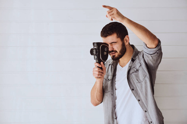 Молодой эмоциональный человек во время съемки со старой камерой на светлом фоне - Фото, изображение