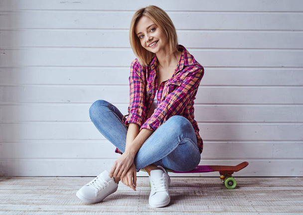 Portrét mladé veselé krásné blondýny dívka sedí na skateboardu a usmívá se na kameru na světlém pozadí - Fotografie, Obrázek