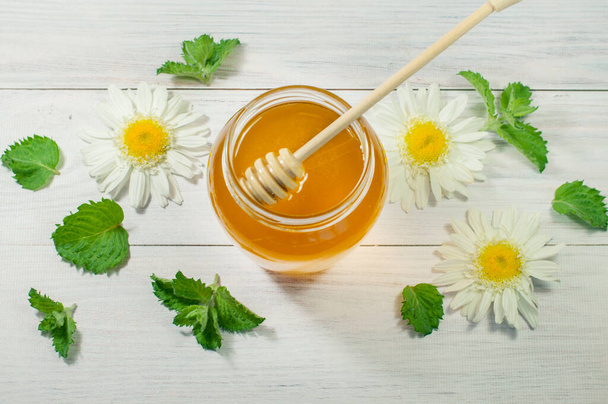 frischer Honig in einem Glas mit einem Löffel steht auf einem hölzernen Hintergrund, Minze und Kamille drumherum - Foto, Bild