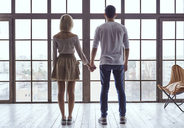 Young happy loving couple guy and girl standing in front of big panoramic window, back to camera holding hands - Foto, Imagem