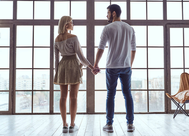 Jeune couple heureux amoureux mec et fille debout devant une grande fenêtre panoramique, retour à la caméra tenant la main - Photo, image