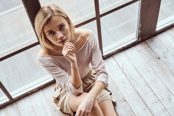 Ritratto di giovane bella ragazza seduta sul pavimento vicino a una grande finestra panoramica e sorridere alla macchina fotografica - Foto, immagini