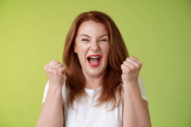 Yeah we did it. Joyful lucky redhead middle-aged female winner pump fists up celebration success gesture yelling triumph joy smiling broadly celebrate awesome news stand green background happy - Photo, Image