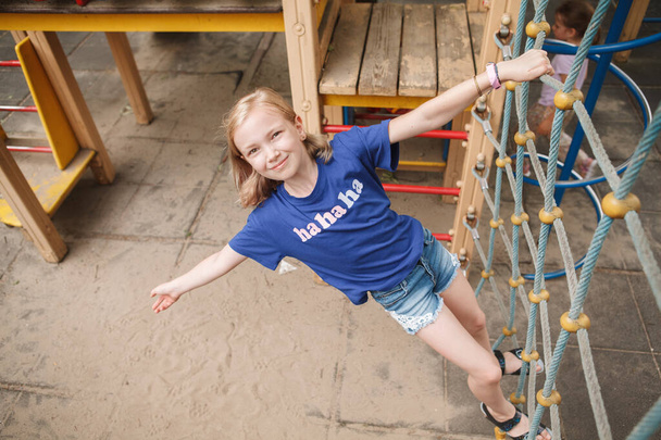 Vista superior de una joven feliz escalando la red de cuerda en el patio de recreo, sonriendo a la cámara - Foto, Imagen