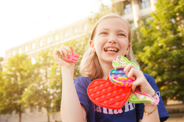 Χαρούμενη νεαρή κοπέλα γελώντας, κοιτάζοντας μακριά, κρατώντας διάφορα πολύχρωμα fidget ποπ παιχνίδια - Φωτογραφία, εικόνα