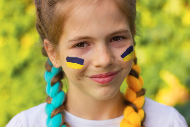 Ein lächelndes ukrainisches Mädchen mit blau-gelben Fahnen auf den Wangen. Patriotismus-Konzept, Flaggen- und Unabhängigkeitstag der Ukraine. Sport und Unterstützung der Fans. - Foto, Bild