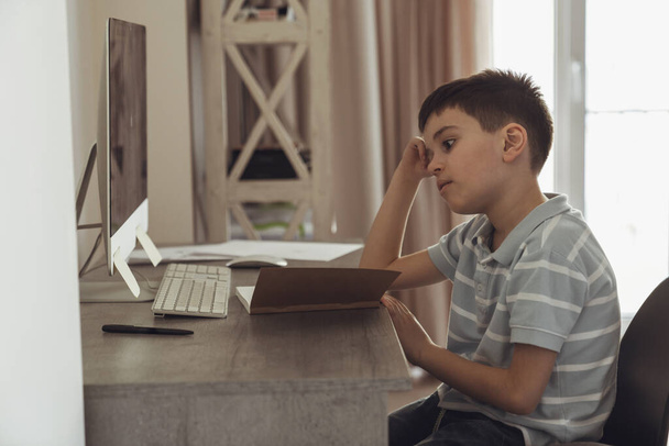 Μικρό αγόρι κάθεται στο δωμάτιό του σε ένα γραφείο δίπλα σε έναν υπολογιστή και κάνει την προετοιμασία για το σχολείο - Φωτογραφία, εικόνα