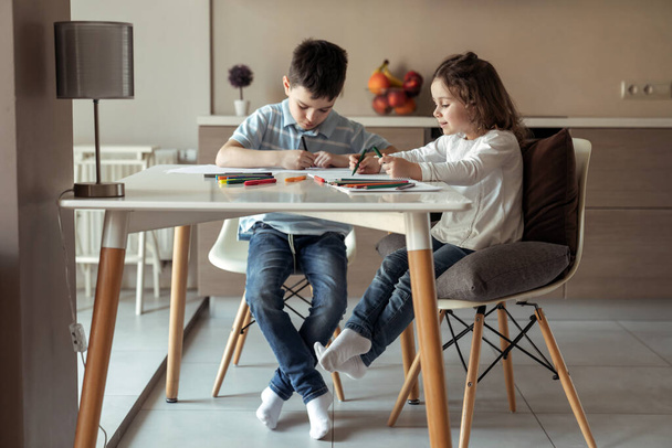 Kleine gelukkige broer en zus zitten aan een tafel thuis in de keuken en tekenen een tekening met gekleurde potloden en stiften - Foto, afbeelding