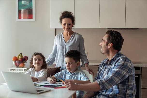 Äiti auttaa pientä tytärtä ja poikaa piirtämään, kun isä työskentelee kotona tietokoneen ja asiakirjojen kanssa, jotka istuvat pöydässä keittiössä ja hymyilevät katsellen vaimoaan kohti. - Valokuva, kuva