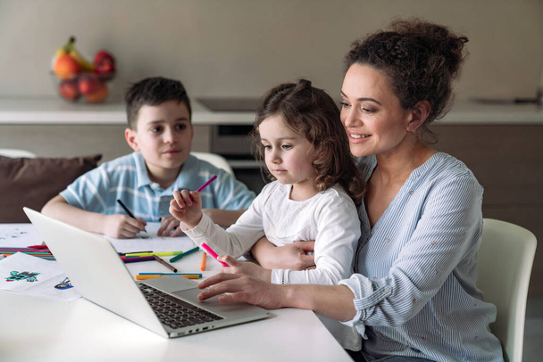 Мама дома с компьютером и документами сидит за столом на кухне и ее маленькая дочь и сын рисует цветными карандашами и маркерами - Фото, изображение