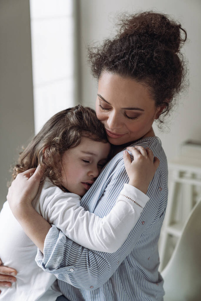 Gute Familie, Mutter und Tochter. Kleine Tochter umarmt ihre Mutter zu Hause mit einem Lächeln und geschlossenen Augen - Foto, Bild