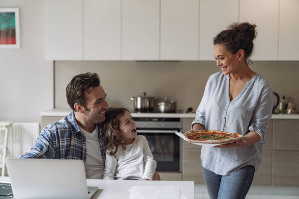 Young woman makes pizza and treats her husband to a freelancer working at home with a computer and documents with her little daughter sitting at a table in the kitchen - Foto, Imagem