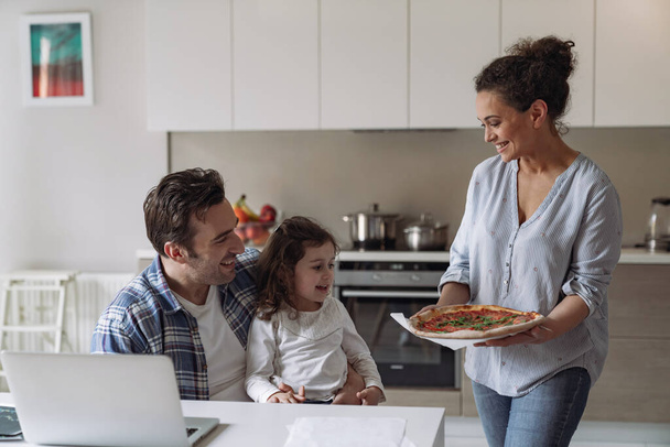 Νεαρή γυναίκα φτιάχνει πίτσα και κερνάει τον άντρα της σε έναν ελεύθερο επαγγελματία που δουλεύει στο σπίτι με έναν υπολογιστή και έγγραφα με τη μικρή της κόρη να κάθεται σε ένα τραπέζι στην κουζίνα - Φωτογραφία, εικόνα
