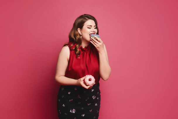 Gorgeous plump woman with bright makeup in modish outfit eating tasty donuts over pink background, isolated with copy space - Photo, image