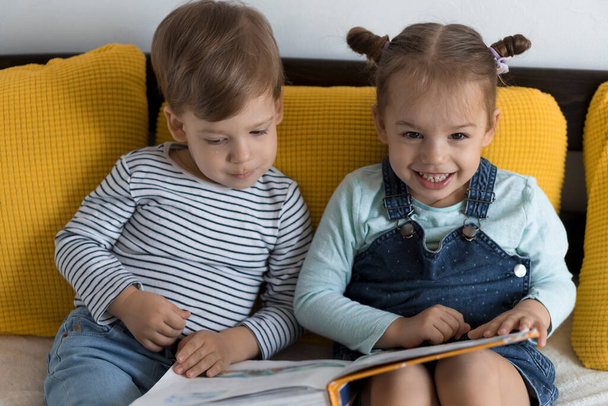 Two preshool, toddler children read large interesting book of fairy tales on yellow bed. Siblings little twins reader have fun, happy kid on quarantine at home. Friendship, family, education concept - Φωτογραφία, εικόνα