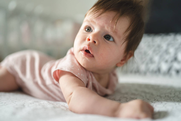 Pieni valkoihoinen vauva neljän kuukauden ikäinen makaa sängyllä kotona valoisassa huoneessa päivällä ja kopioi tilaa katsomalla sivumahaan. - Valokuva, kuva