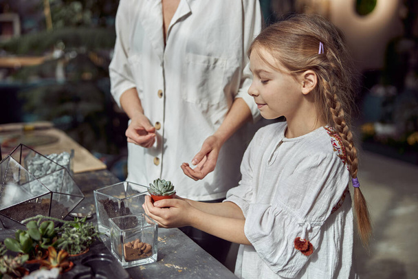Kaukasische selbstbewusste, glückliche Floristin arbeitet mit ihrer kleinen Tochter und fertigt im Botanischen Geschäft Kompositionen aus Glassteinen und Pflanzen - Foto, Bild