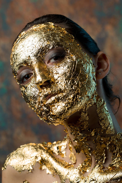 Girl with a mask on her face made of gold leaf. Gloomy studio portrait of a brunette on an abstract background. - Valokuva, kuva