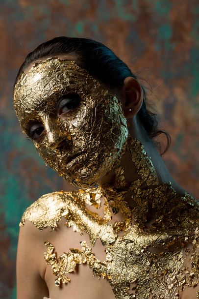 Girl with a mask on her face made of gold leaf. Gloomy studio portrait of a brunette on an abstract background. - Fotografie, Obrázek