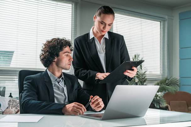 Businesswoman and businessman in formal suit is working together pensively looking into a black folder. Laptop on desk. Office background. Concept of teamwork - Fotografie, Obrázek