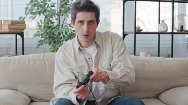 Caucasico millennial adulti 30s focalizzato uomo maschio giocatore ragazzo seduto a casa sul divano in salotto interno godendo concorrenza video gioco console controlli joystick gioco - Filmati, video