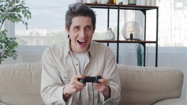 Portré érzelmi felnőtt koncentrált kaukázusi férfi millenniumi férfi gamer srác otthon ül a kanapén videojáték konzol online izgatott a verseny számítógépes függőség élvezi a szerencsejáték - Felvétel, videó