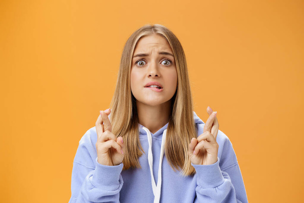 Worried anxious hopeful young woman with fair hair in stylish over-sized hoodie biting lower lip frowning concerned crossing fingers for good luck praying for dream come true against orange wall - Photo, Image