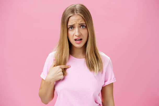 Insulted girl pointing at herself with displeased pissed and questioned expression asking question being shocked she picked or accused in something terrible and disrespectful posing against pink wall - 写真・画像