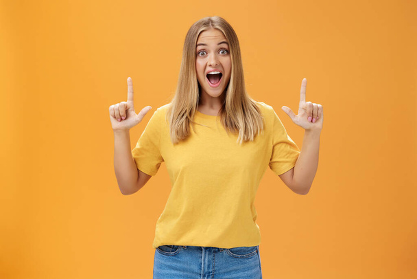 Portrait of joyful surprised and impressed delighted young woman with tanned skin and fair hair opening mouth from amazement and joy raising hands pointing up at awesome copy space against orange wall - Zdjęcie, obraz