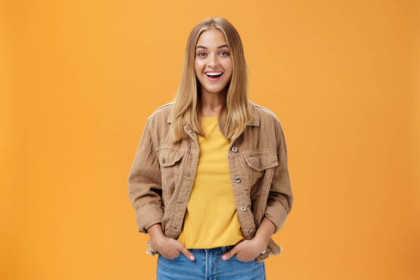 Charismatic tanned woman in corduroy jacket and yellow t-shirt ready for chilly autumn walk with friends smiling joyfully gazing entertained at camera holding hand in pockets casually over orange wall - Foto, afbeelding