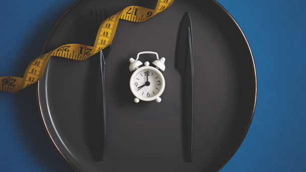 Flat lay fork and kitchen knife on a plate with tape measurement and white alarm clock. Intermittent fasting concept dieting healthy lifestyle - Photo, Image